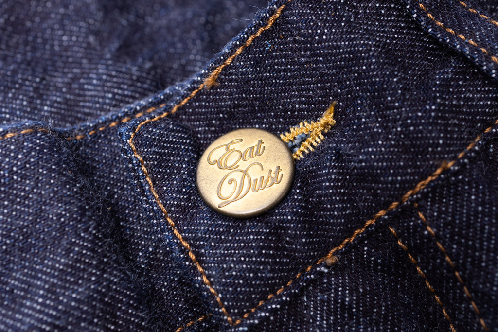 Close-up of a gold 'Eat Dust' button on blue denim fabric.