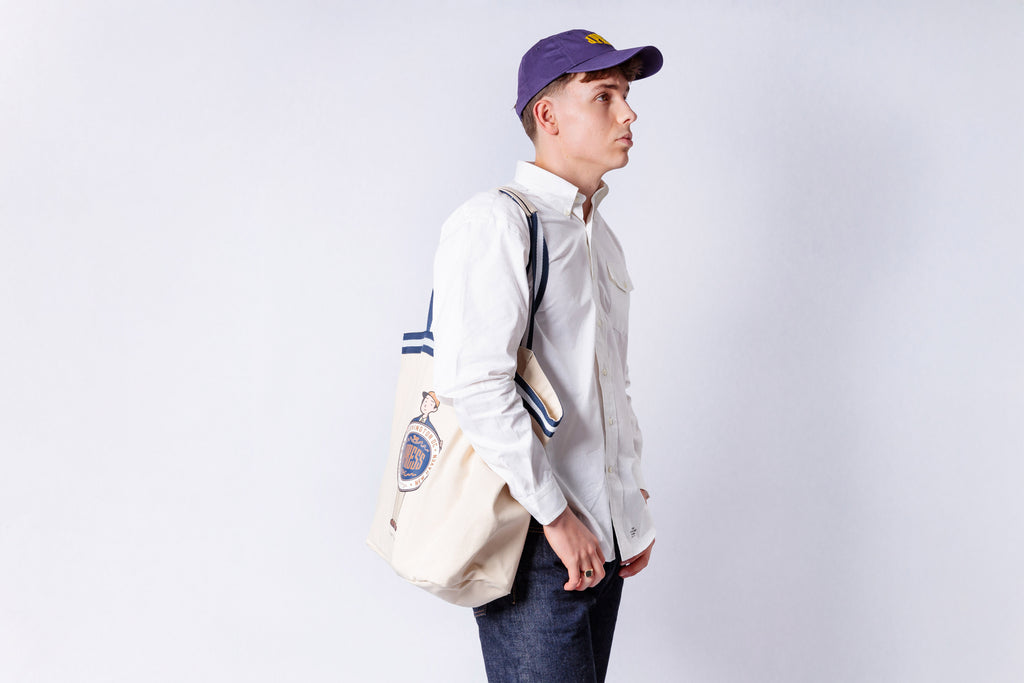 Person wearing a white shirt, blue jeans, and a purple cap with a beige backpack on a light gray background