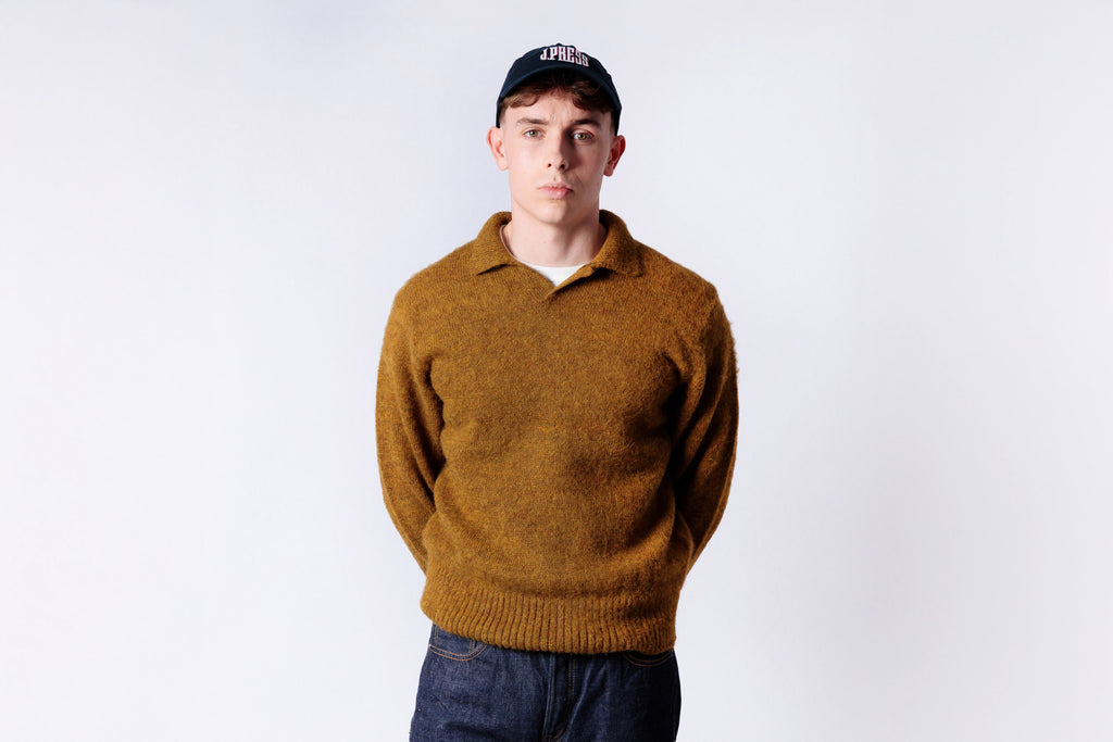 Person wearing a mustard yellow sweater and dark cap on a white background