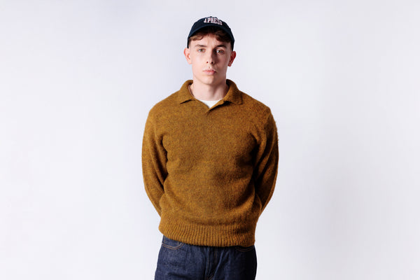 Person wearing a mustard yellow sweater and dark cap on a white background