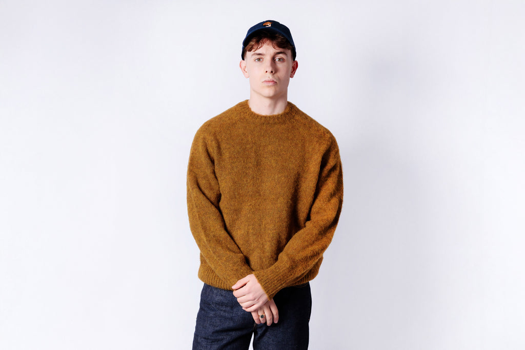 Person wearing a mustard yellow sweater and dark cap on a white background