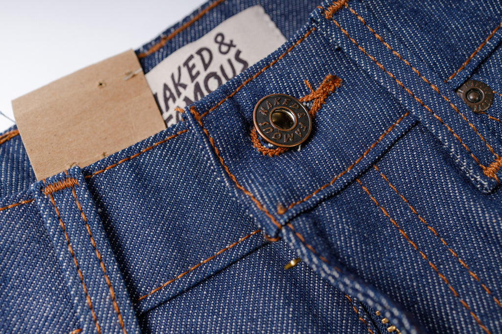 Close-up of blue denim fabric with a button and 'Naked & Famous' label.