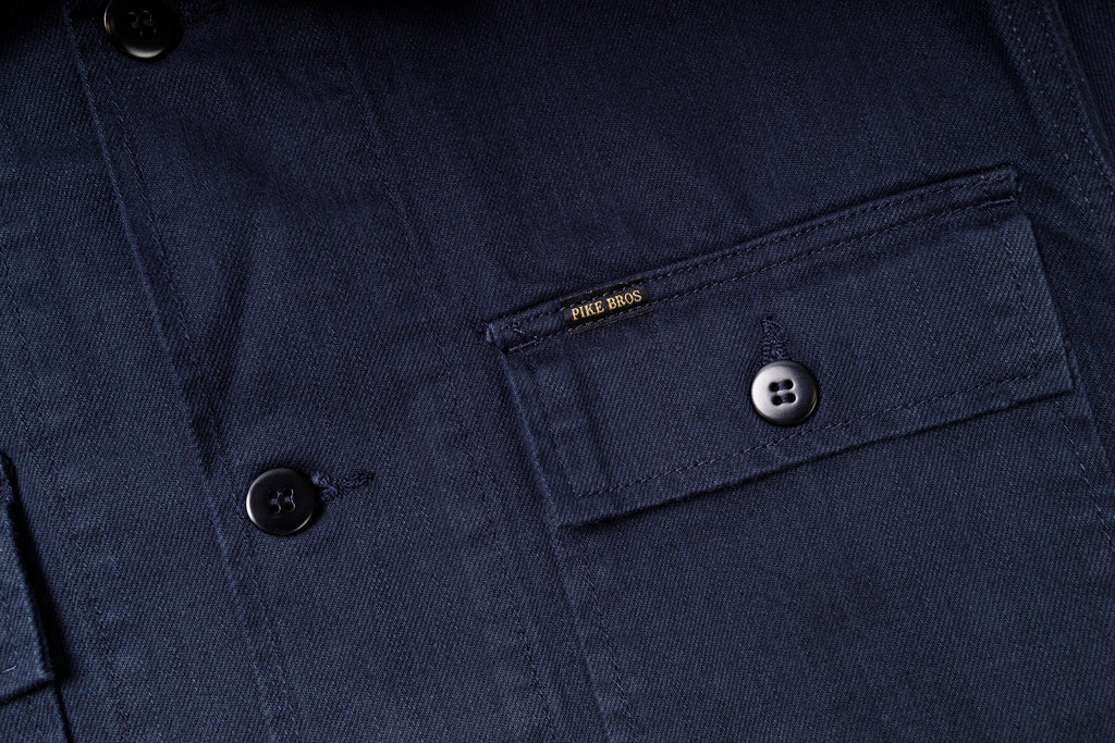 Close-up of a navy blue jacket with a button and pocket detail, featuring the brand 'Pike Bros'.