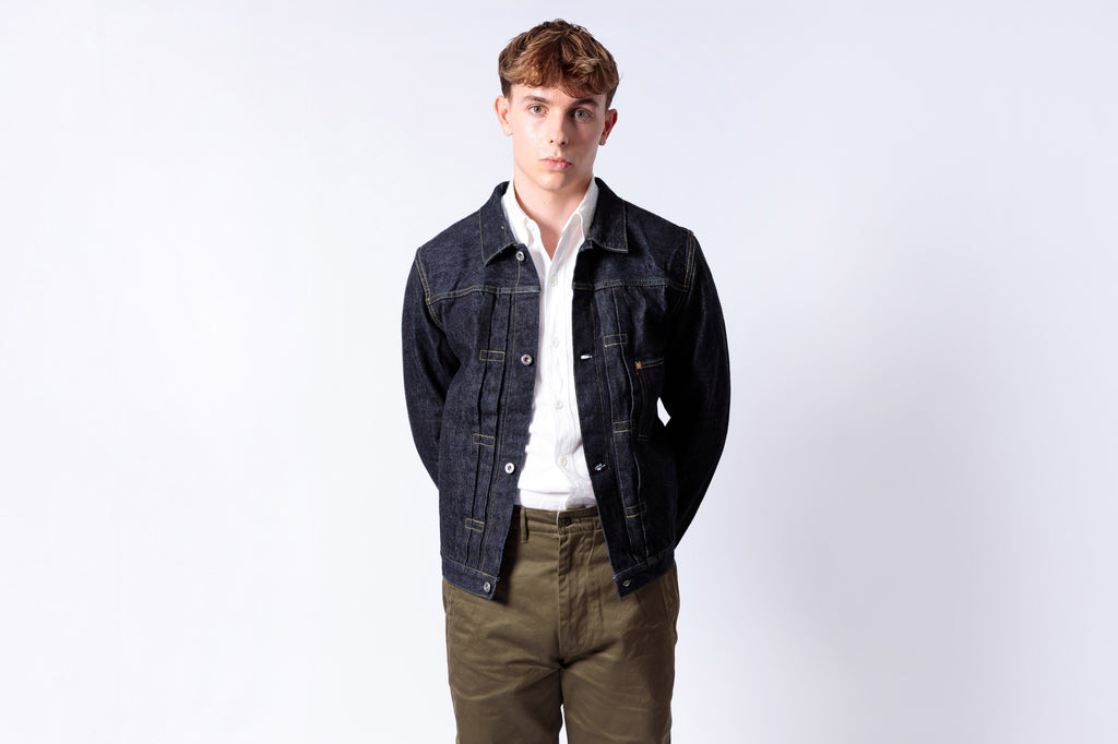 Man wearing a dark denim jacket over a white shirt and green pants on a white background