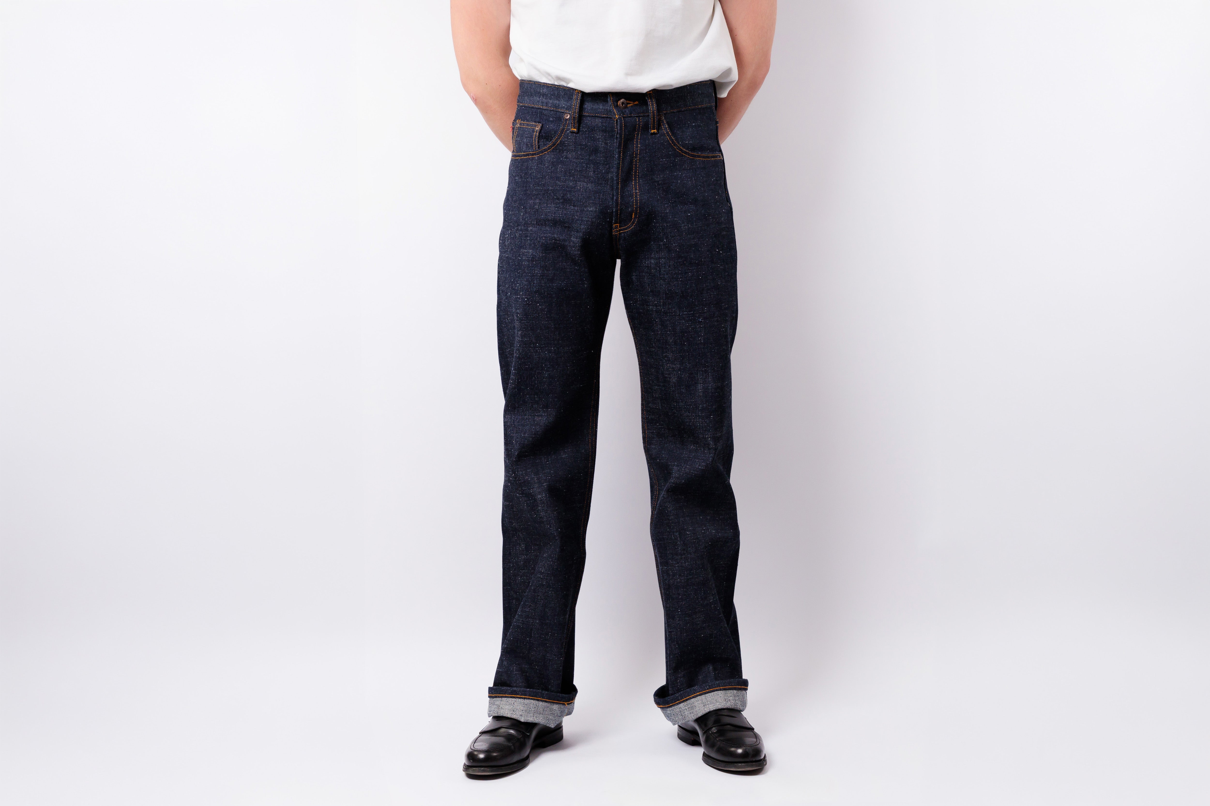 A person wearing dark indigo blue straight-leg jeans, a white t-shirt, and black shoes, standing against a light gray studio background.