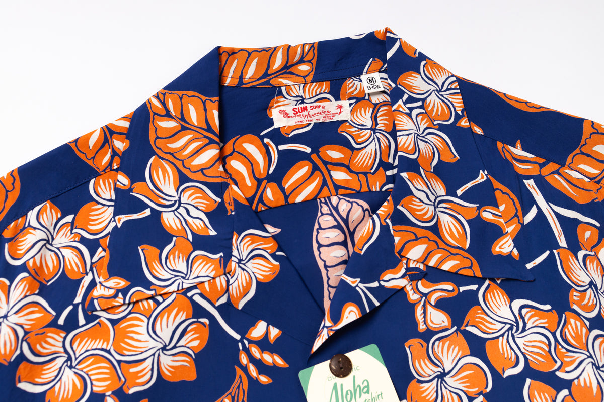 A close-up of a vintage Hawaiian shirt featuring a bold orange and white hibiscus flower and leaf pattern on a navy blue background. 