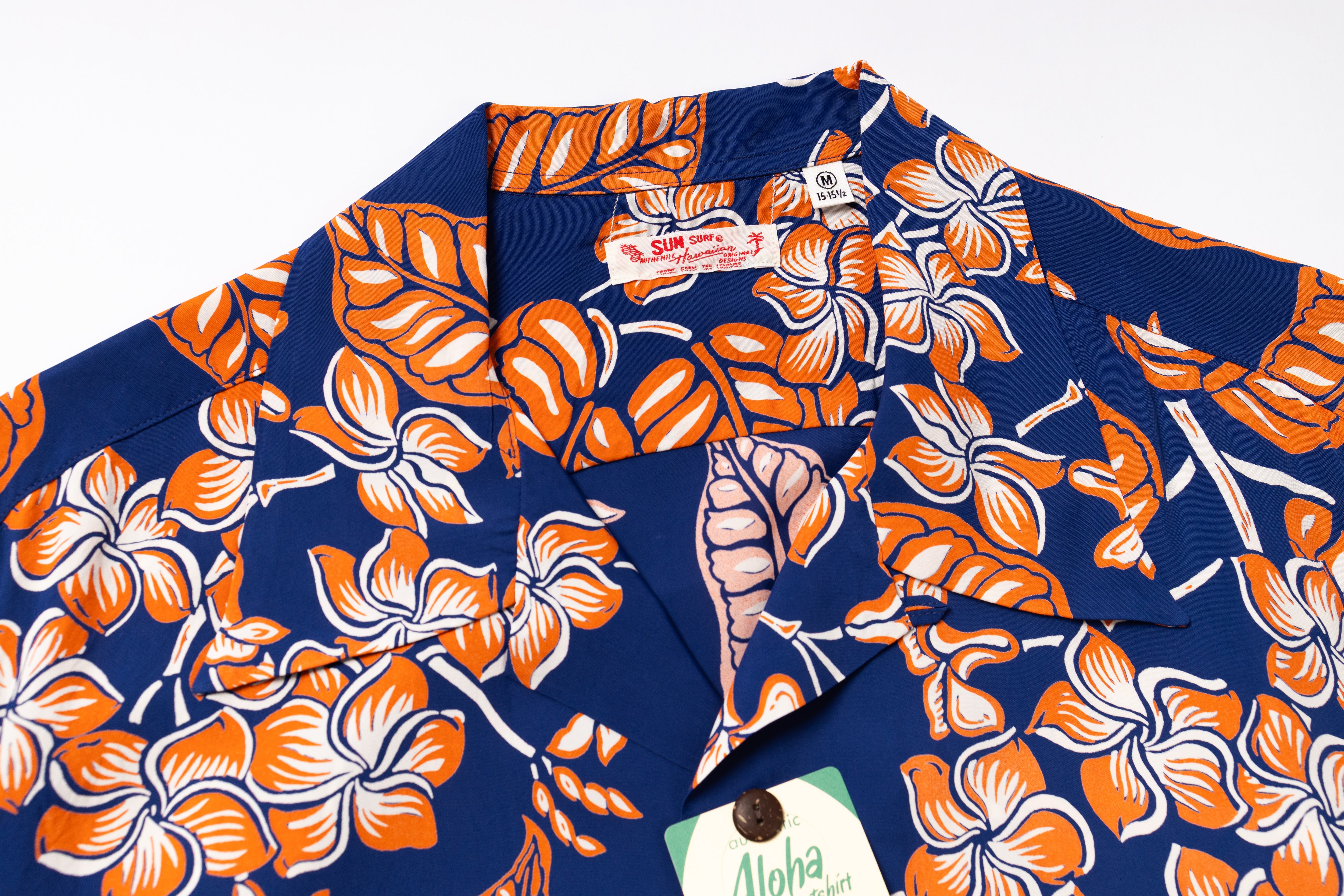 A close-up of a vintage Hawaiian shirt featuring a bold orange and white hibiscus flower and leaf pattern on a navy blue background. 