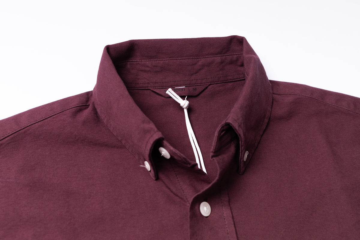 A close-up of a button-down shirt collar in a deep burgundy or maroon fabric, with white buttons and a white drawstring tie inside the collar, shown against a plain white background.