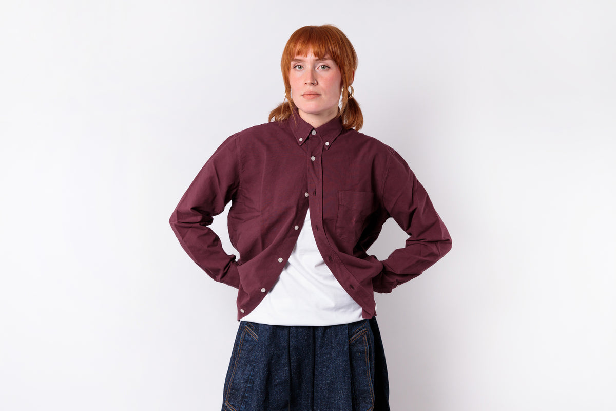 A woman with red hair in pigtails stands against a white studio background wearing a dark burgundy button-up overshirt over a white t-shirt and dark blue denim skirt.