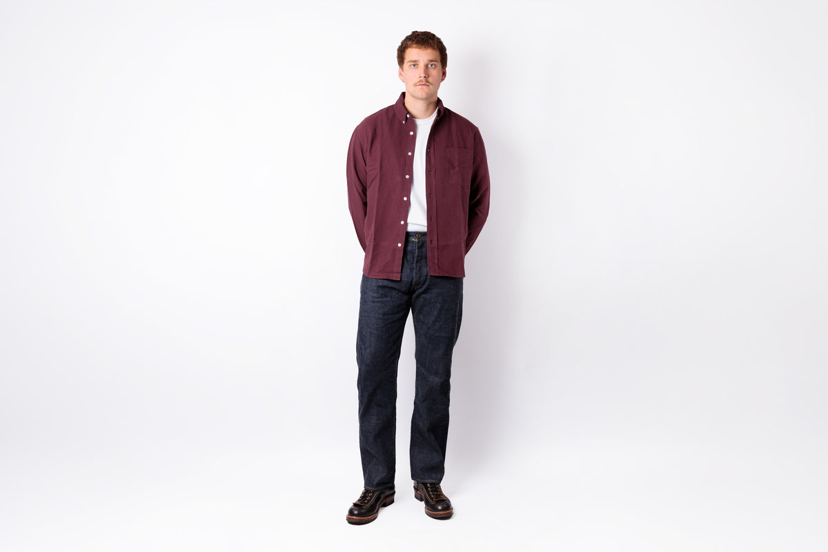A man stands against a plain white studio background wearing a burgundy corduroy button-up shirt jacket over a white t-shirt, dark blue jeans, and brown shoes.