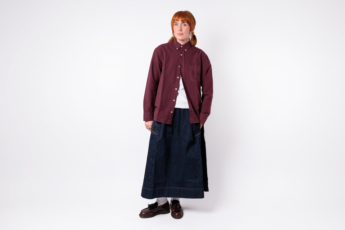 A model stands against a plain white studio background wearing a dark burgundy button-up jacket over a white top, paired with a long dark navy skirt and dark brown shoes.