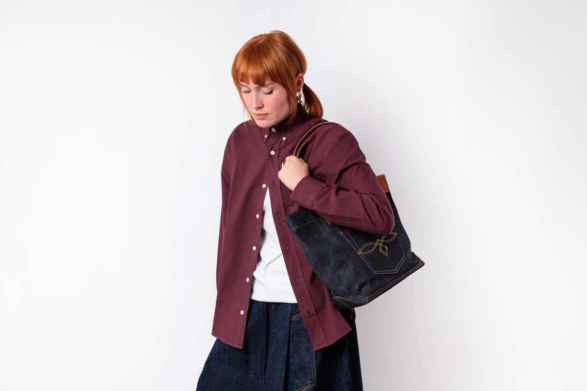 A woman with red hair tied back wears a burgundy button-up shirt over a white top and dark denim jeans, carrying a dark denim tote bag with brown leather straps and gold-toned stitching against a plain white background.