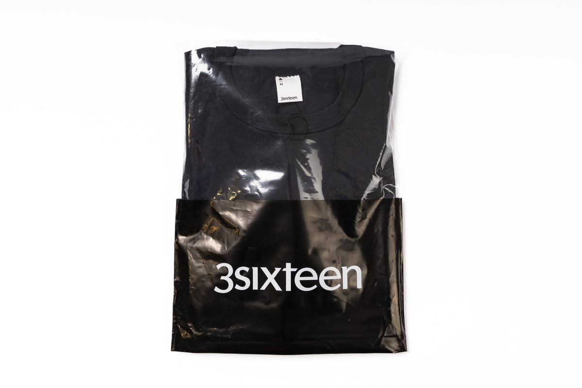 Black t-shirt in a clear plastic bag with '3sixteen' branding on a white background