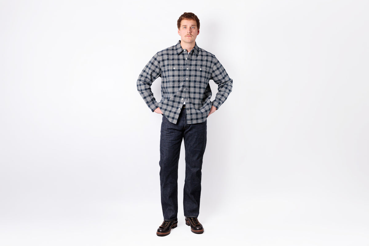 A man stands against a white studio background wearing a long-sleeve gray and black plaid button-down shirt, dark blue jeans, and brown shoes.