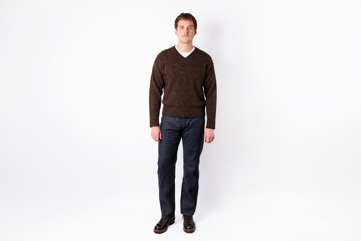 A man wearing a dark brown v-neck knit sweater, dark blue jeans, and brown leather shoes, standing against a plain white studio background.