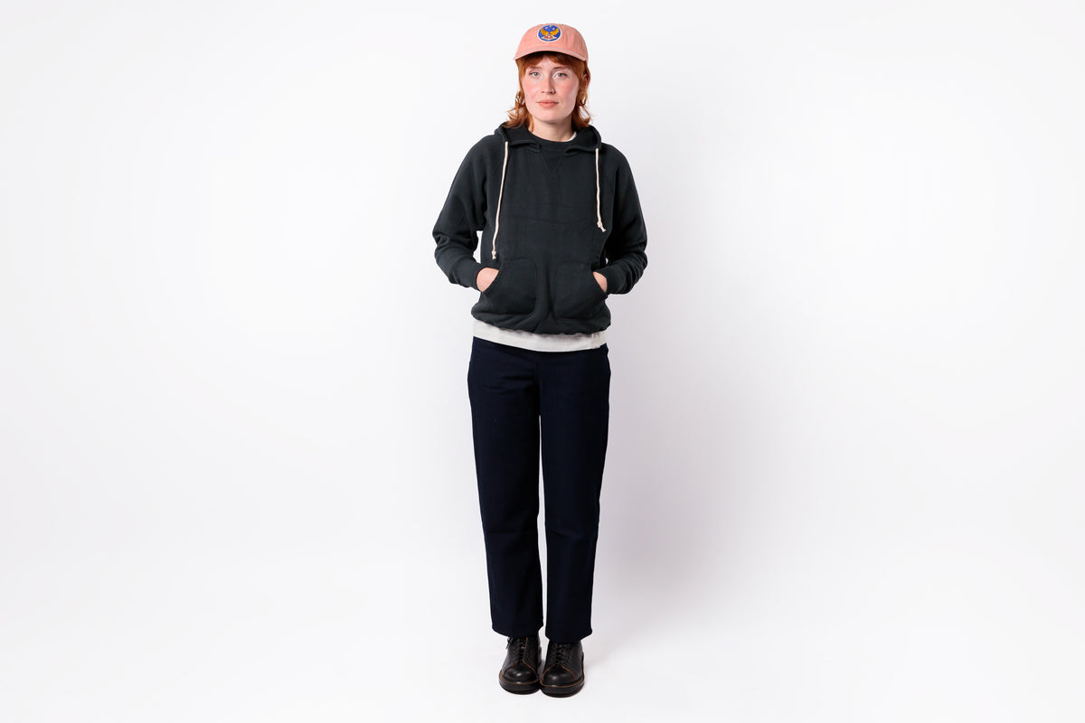 A person stands against a plain white studio background wearing a dark charcoal hoodie with white drawstrings, a light gray shirt underneath, dark navy pants, black shoes, and a salmon-pink baseball cap.