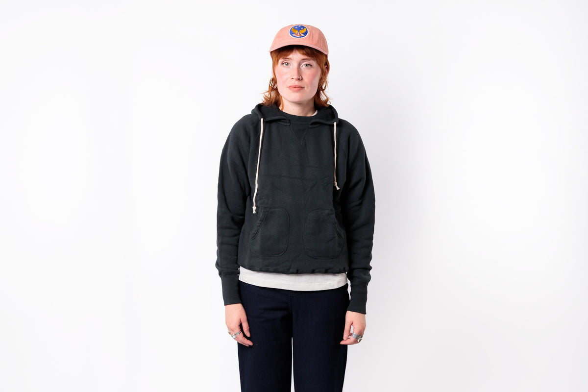 A person standing against a plain white background wearing a dark charcoal gray cropped hoodie with white drawstrings, a light pink baseball cap, a white undershirt, and dark pants.