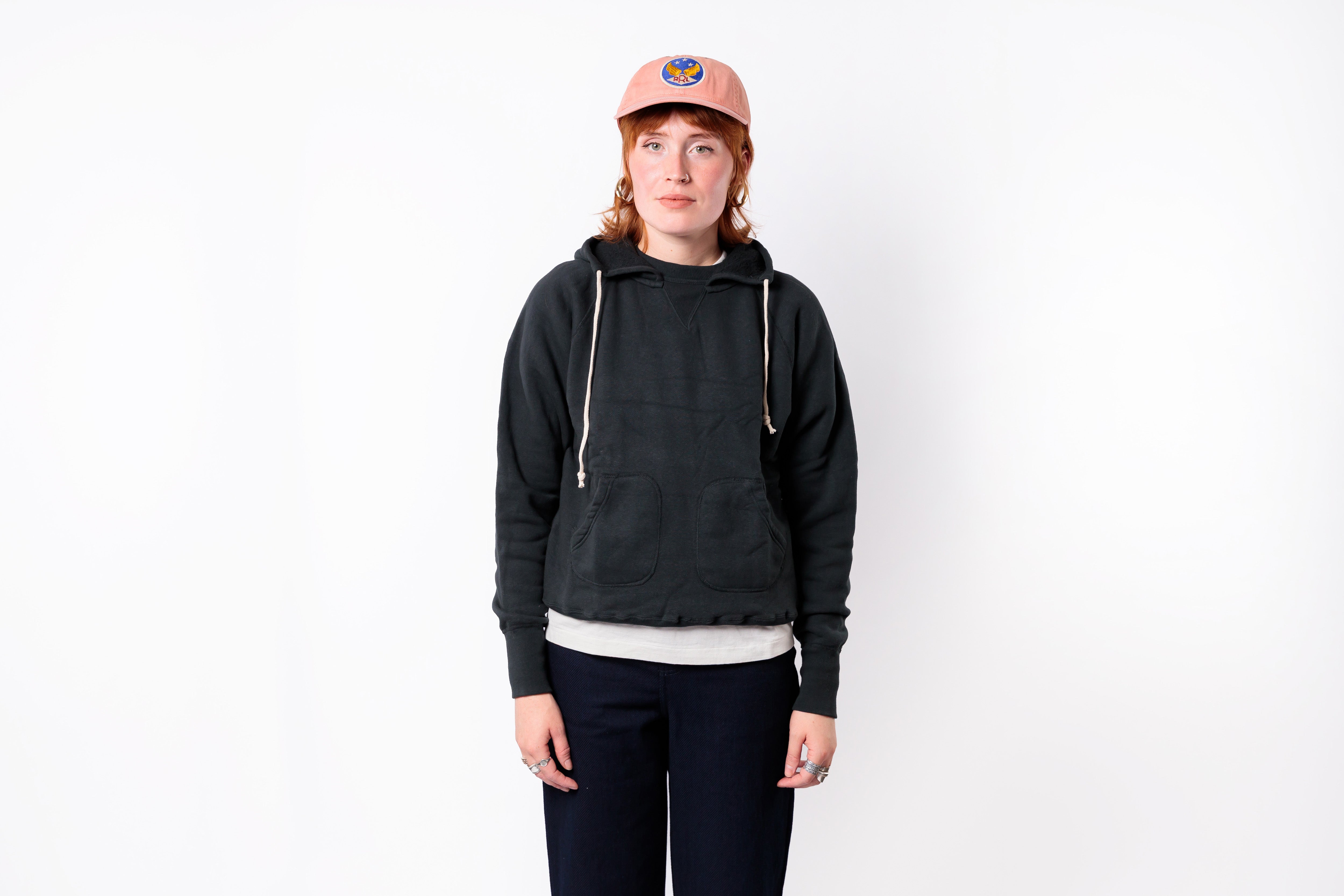 A person standing against a plain white background wearing a dark charcoal gray cropped hoodie with white drawstrings, a light pink baseball cap, a white undershirt, and dark pants.