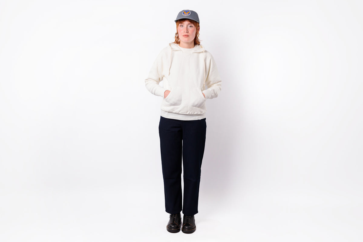 A person standing against a plain white studio background wearing a cream pullover hoodie, dark navy pants, black shoes, and a gray baseball cap.