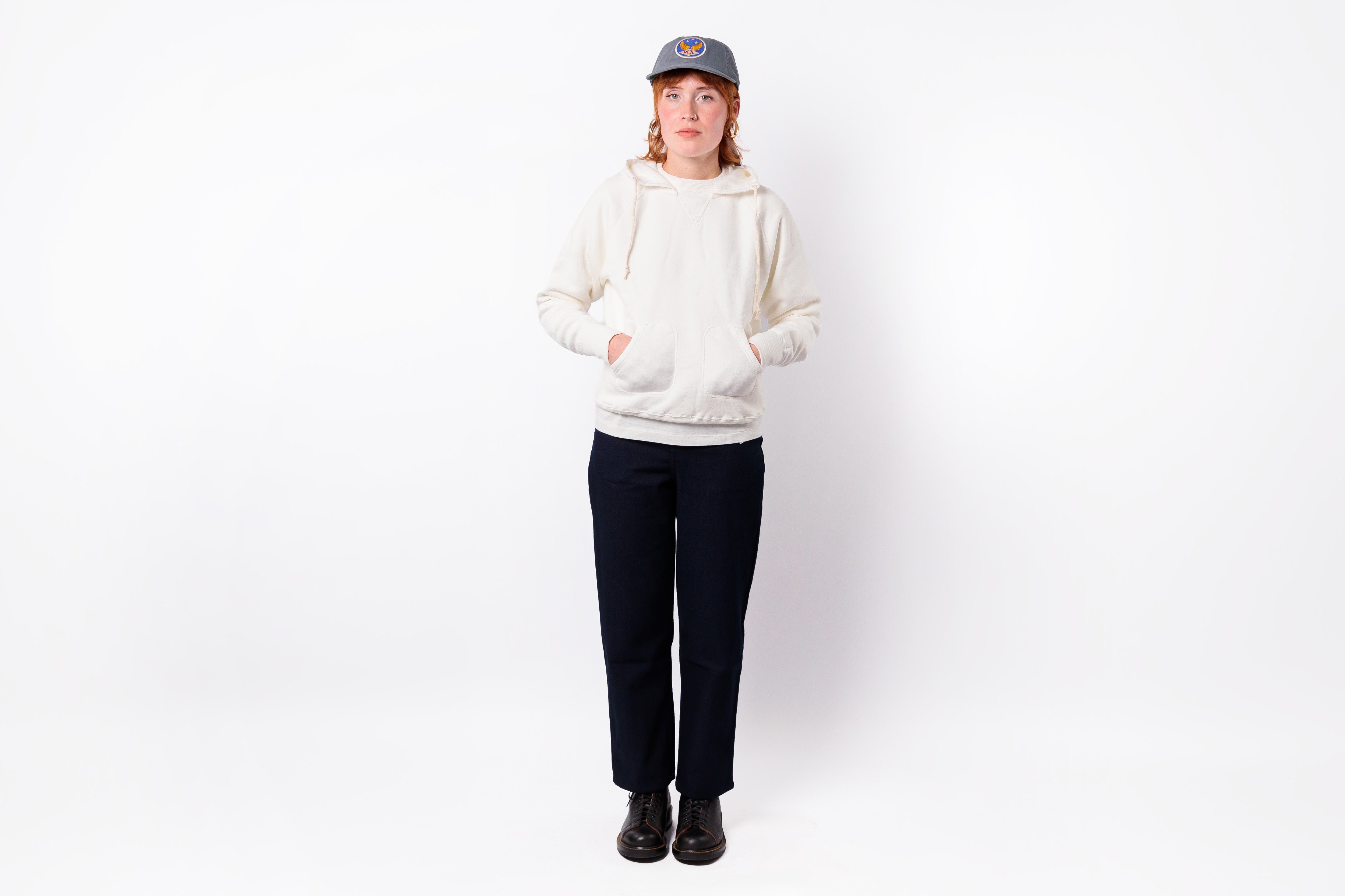 A person standing against a plain white studio background wearing a cream pullover hoodie, dark navy pants, black shoes, and a gray baseball cap.