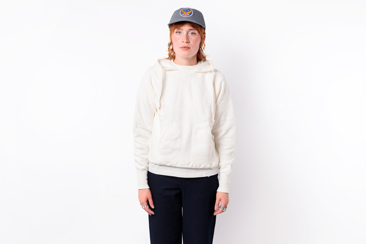 A person standing against a plain white background wearing a cream-colored pullover hoodie, dark navy pants, and a gray baseball cap with a circular logo.