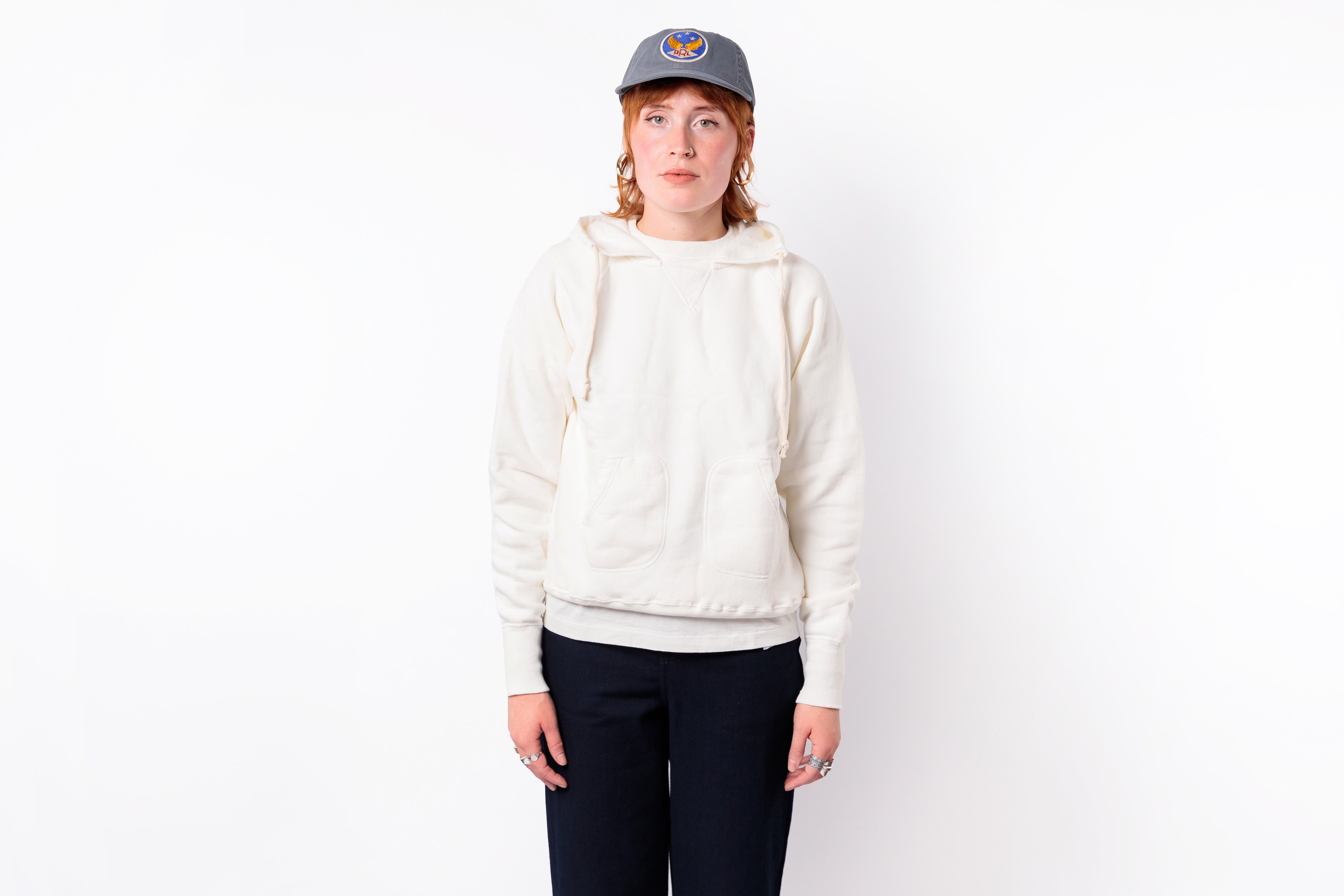 A person standing against a plain white background wearing a cream-colored pullover hoodie, dark navy pants, and a gray baseball cap with a circular logo.