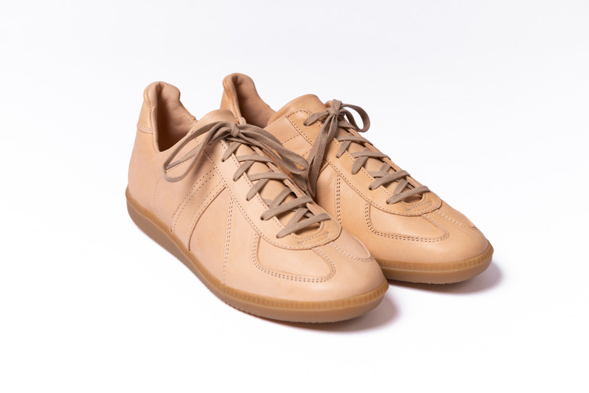 A pair of low-top leather sneakers in a warm tan/beige color, featuring matching beige laces and a gum sole, photographed on a clean white background.