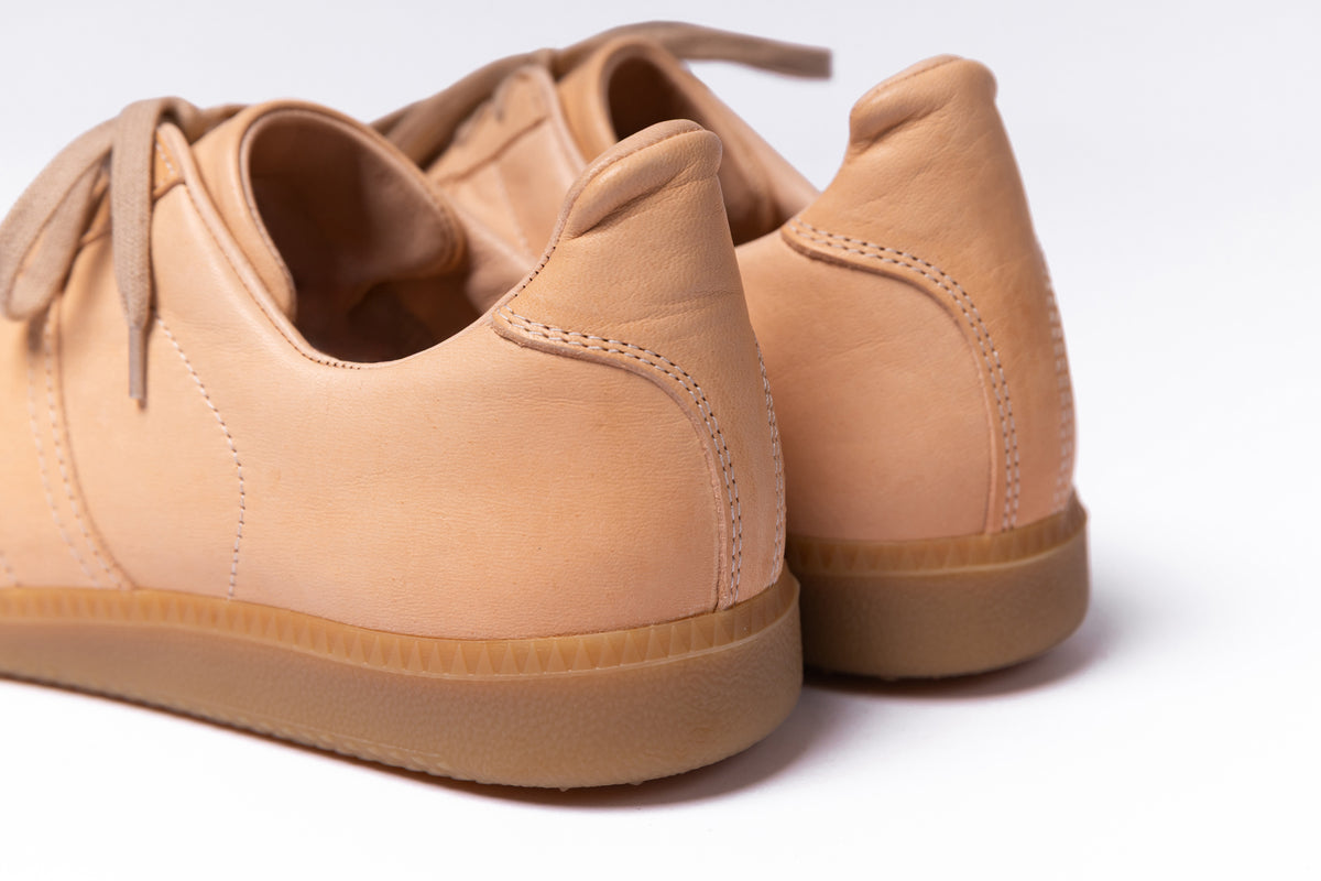 Close-up of a pair of tan leather low-top sneakers with gum soles, shown from the rear three-quarter angle on a white background.