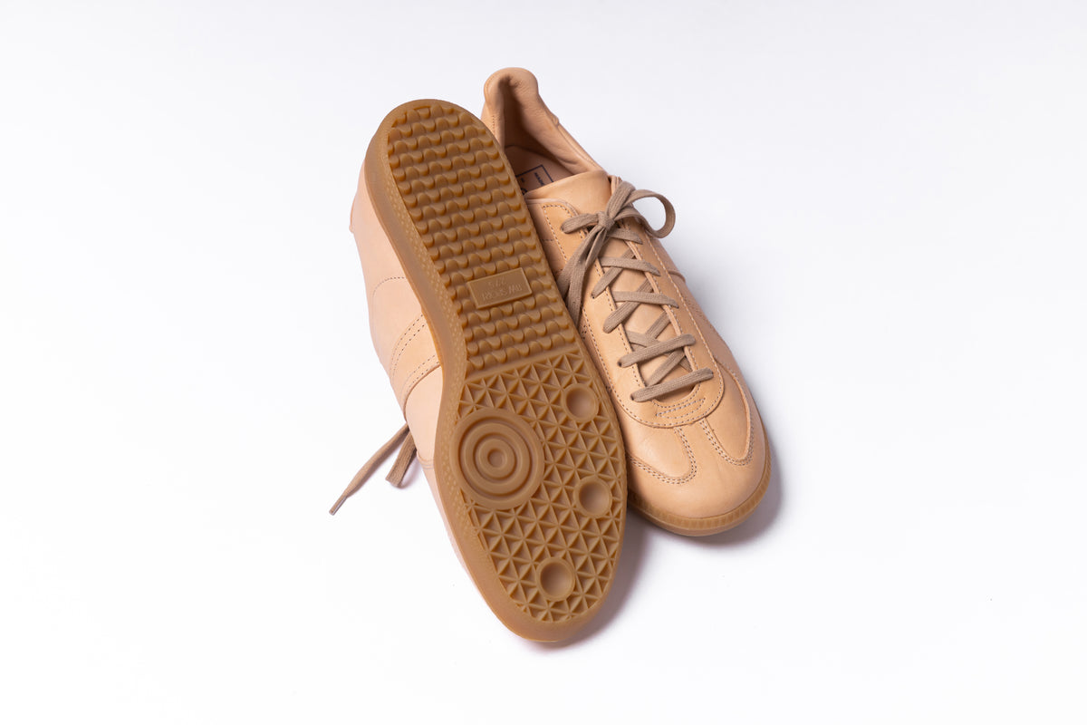 A pair of low-top sneakers in a monochromatic tan colorway, with one shoe showing the textured rubber outsole and the other showing the side profile against a clean white background.