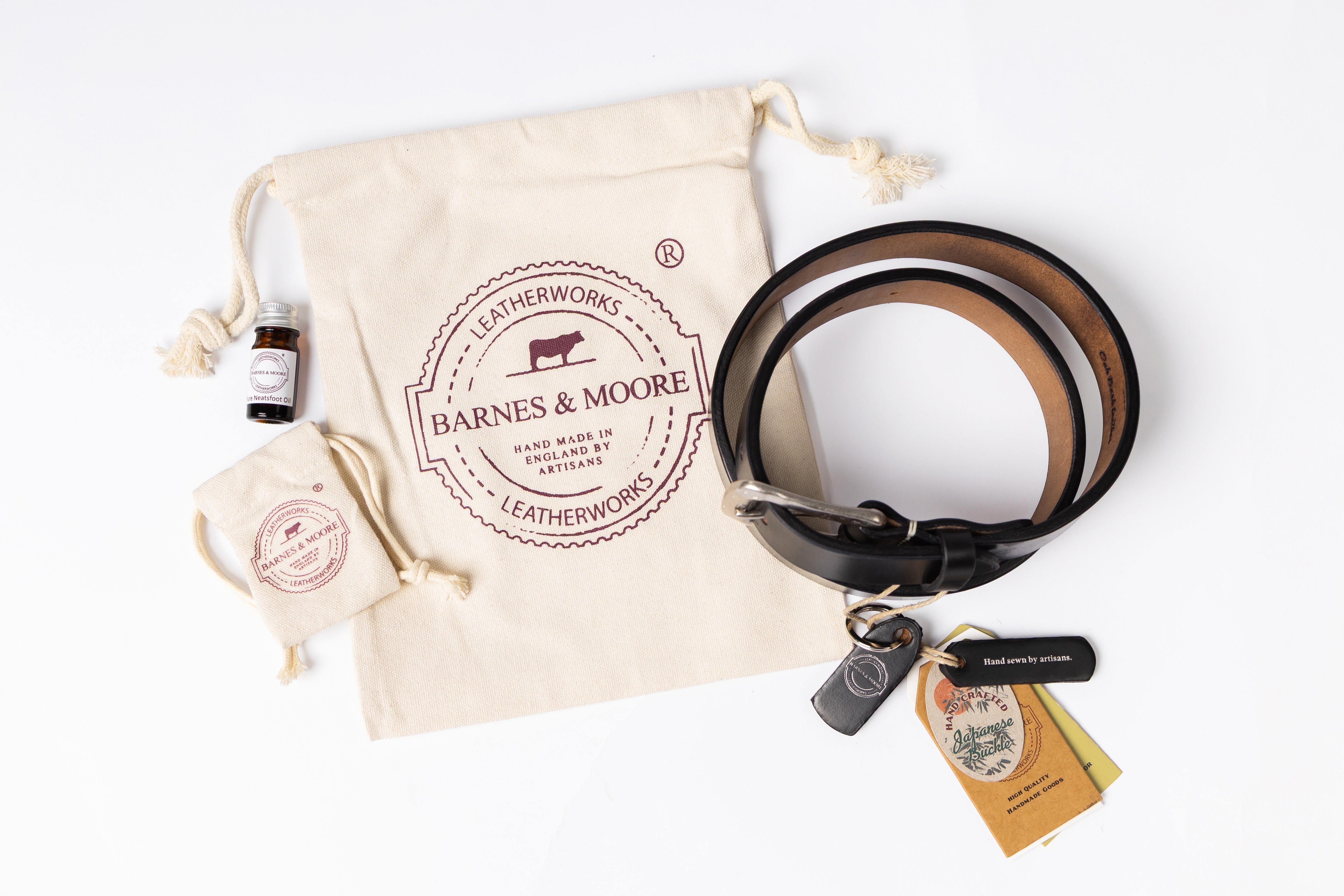 A leather accessory gift set from Barnes & Moore Leatherworks arranged on a white background, including a black leather belt with tan interior, a cream drawstring pouch with the brand logo, a small bottle, a small pouch, and a keychain with tags.