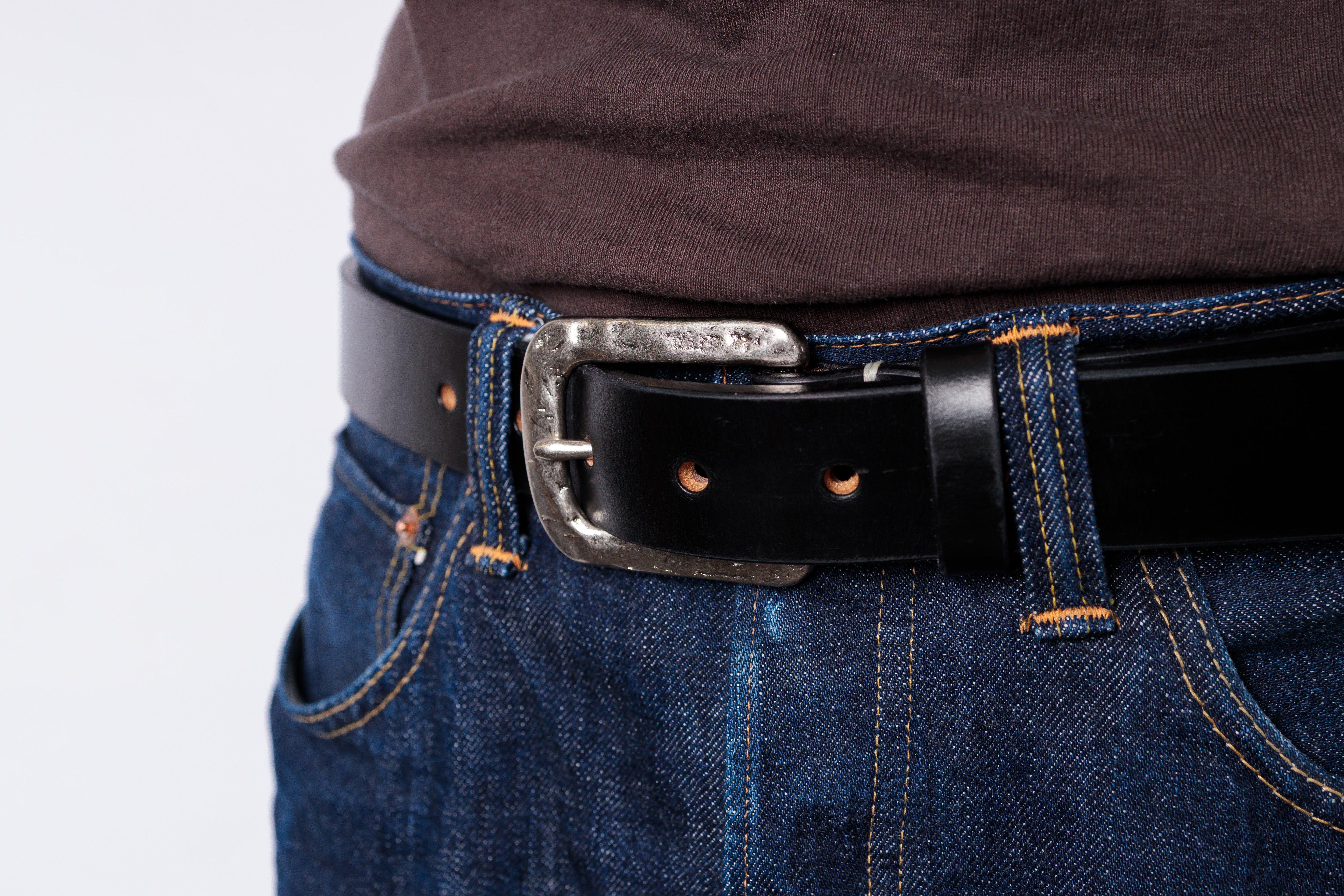 A close-up shot of a black leather belt with a silver-toned metal buckle, worn over a brown shirt and blue jeans.