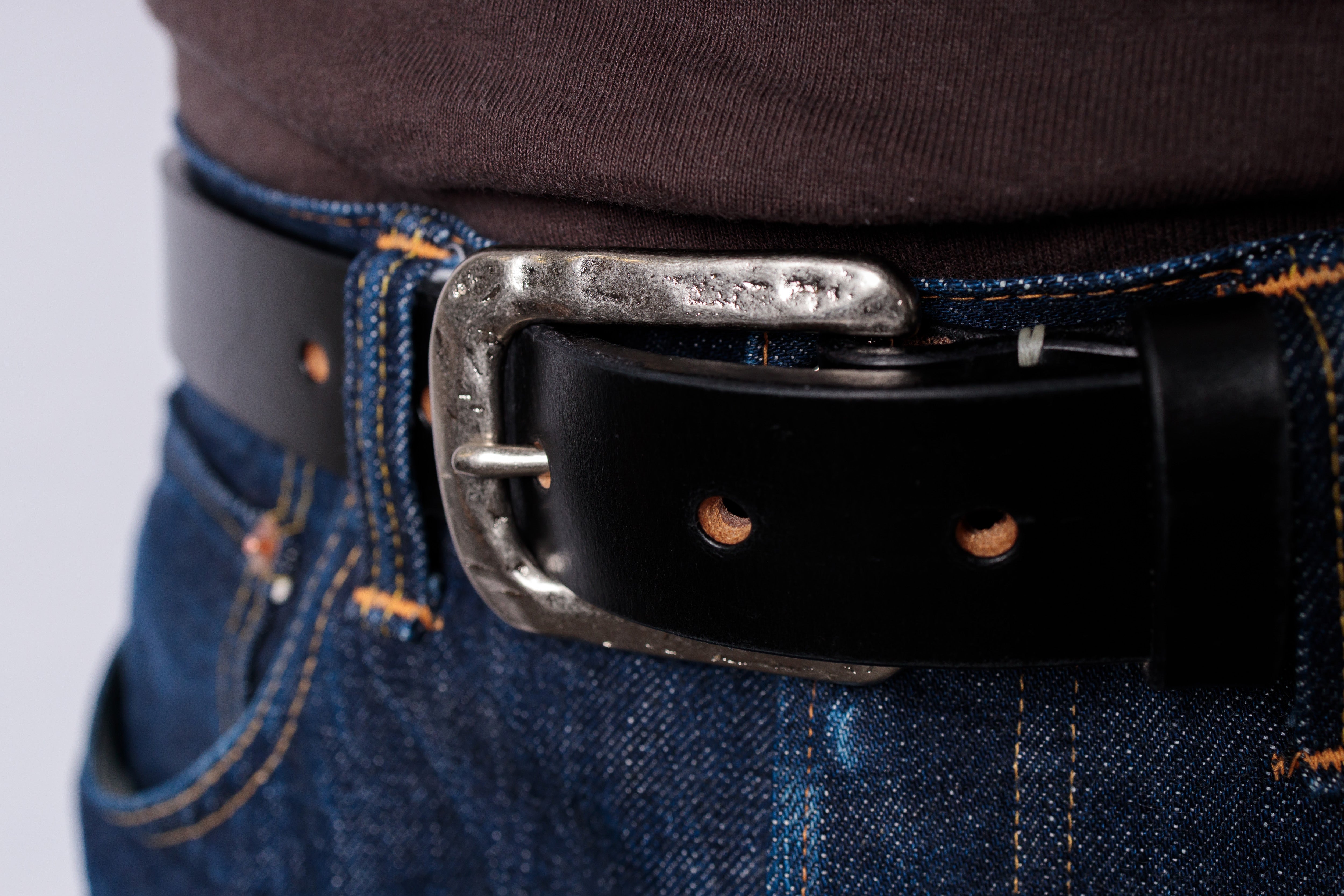 A close-up shot of a black leather belt with a textured silver buckle, worn over blue denim jeans and a dark brown shirt.