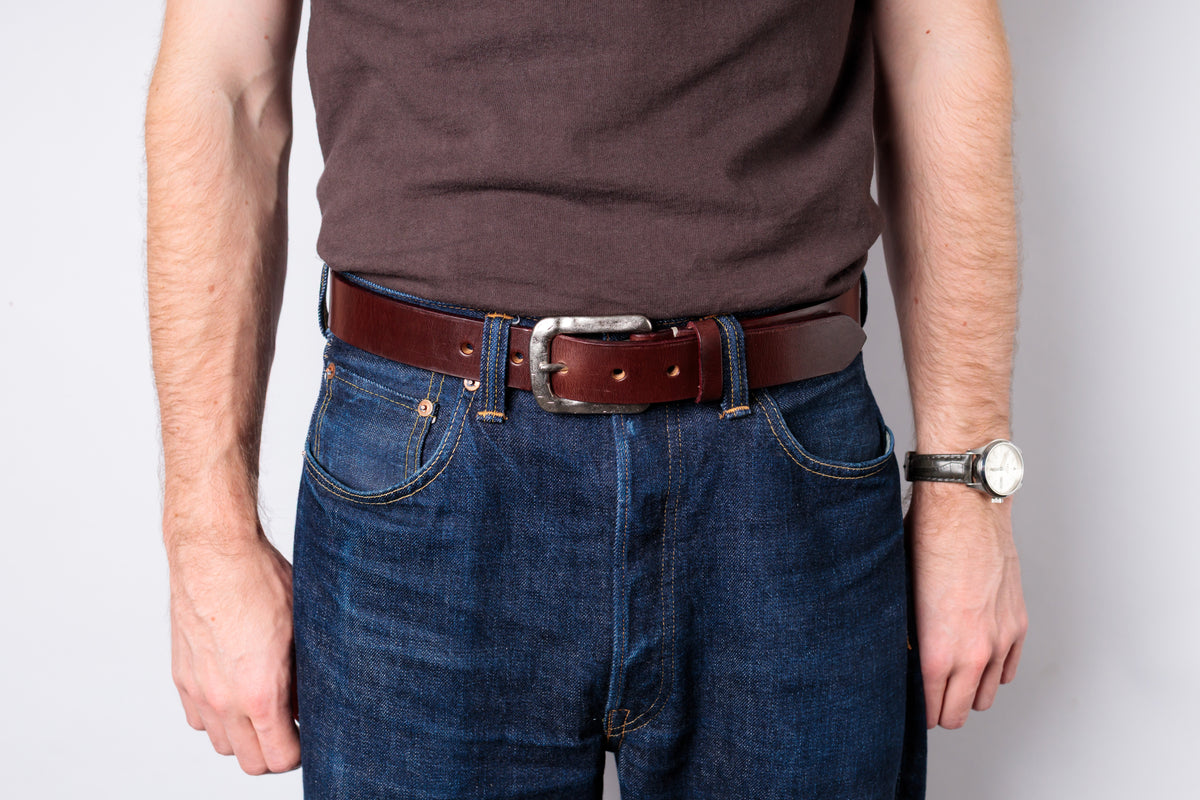 A man wearing a brown leather belt with a silver buckle, styled with dark blue jeans and a brown t-shirt against a light gray background.