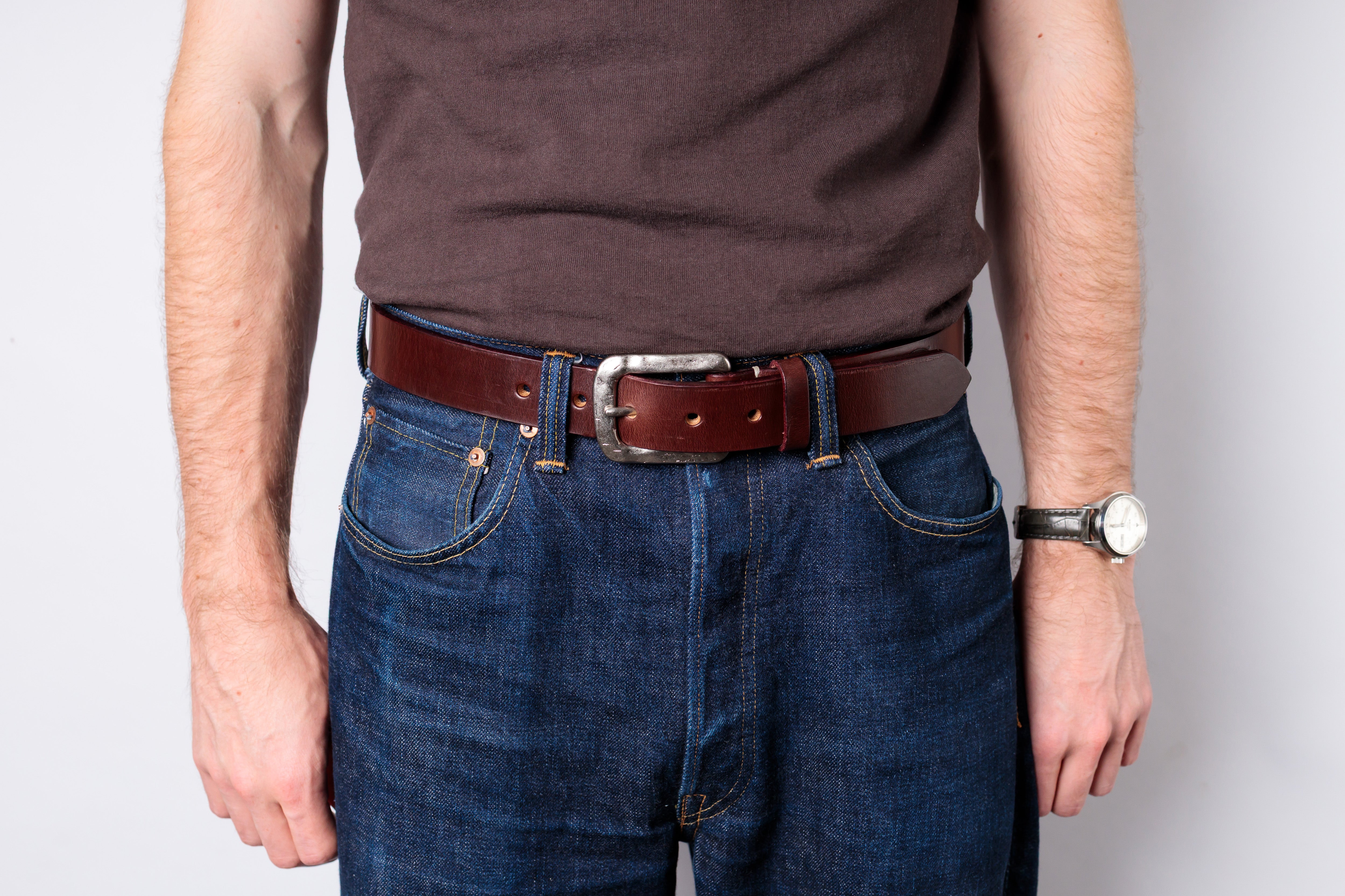 A man wearing a brown leather belt with a silver buckle, styled with dark blue jeans and a brown t-shirt against a light gray background.
