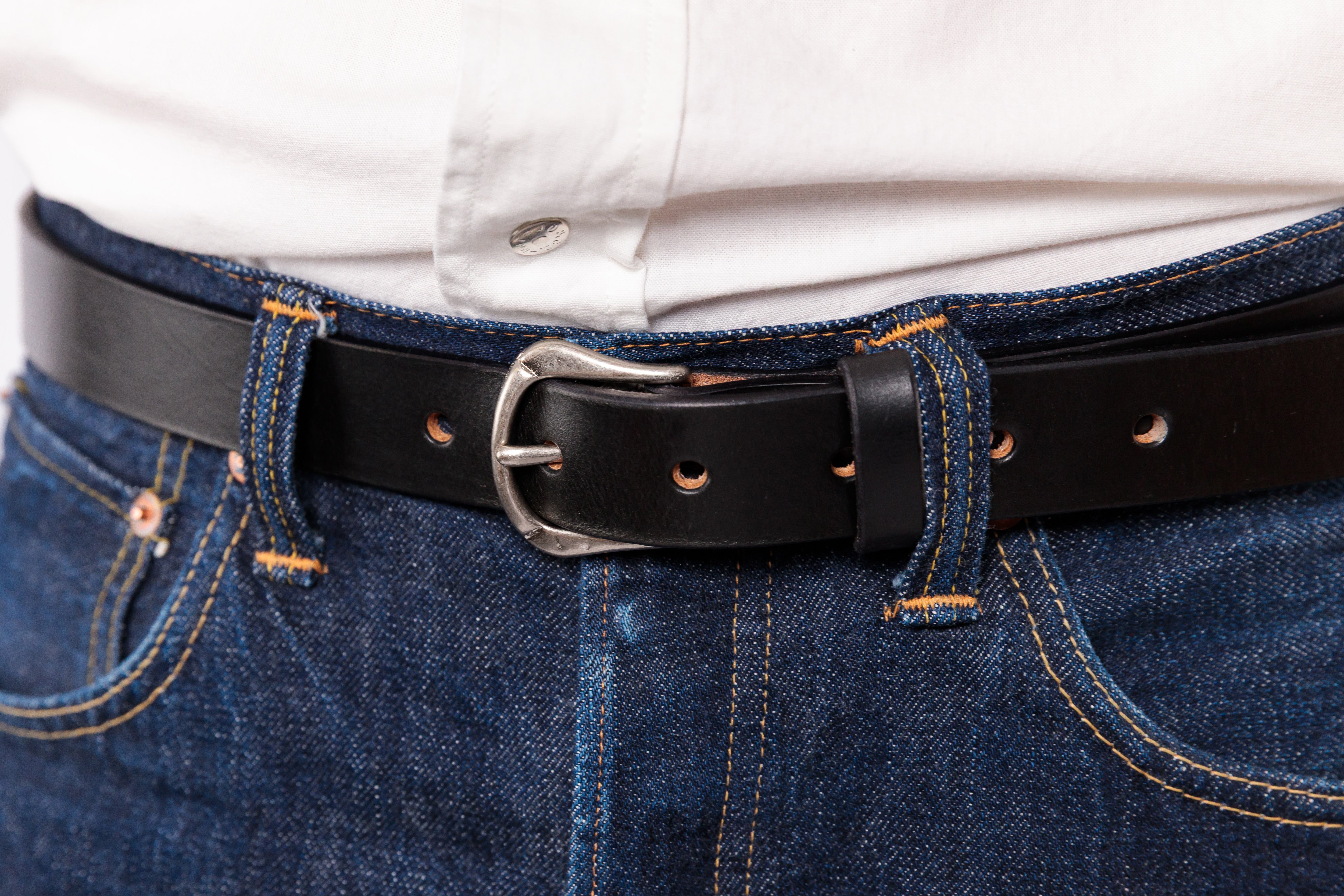 A close-up shot of a black leather belt with a silver buckle worn over blue denim jeans and a white button-down shirt.