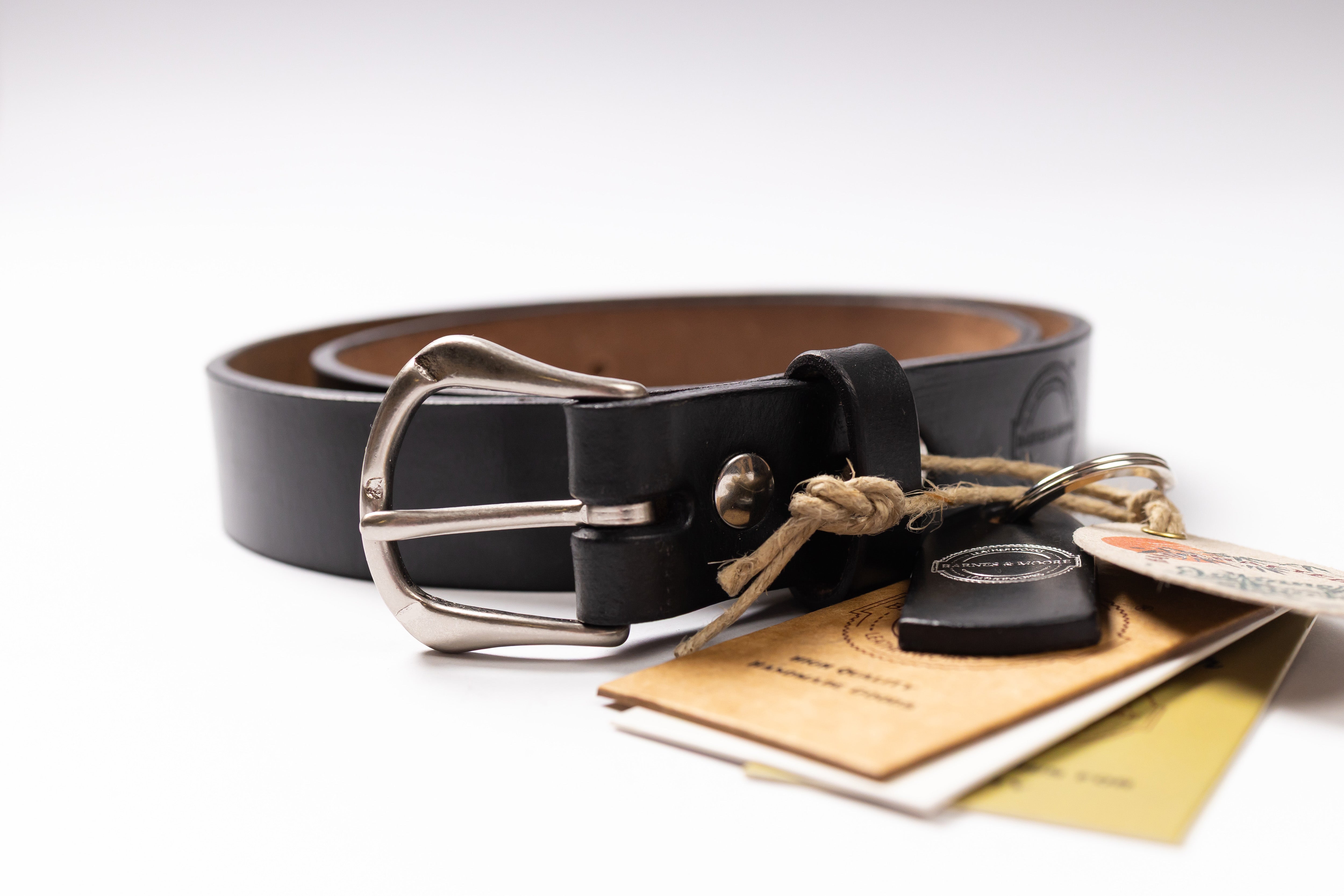 A black leather belt with a silver-tone metal buckle, shown coiled on a white background with its paper tags and a small key fob attached.
