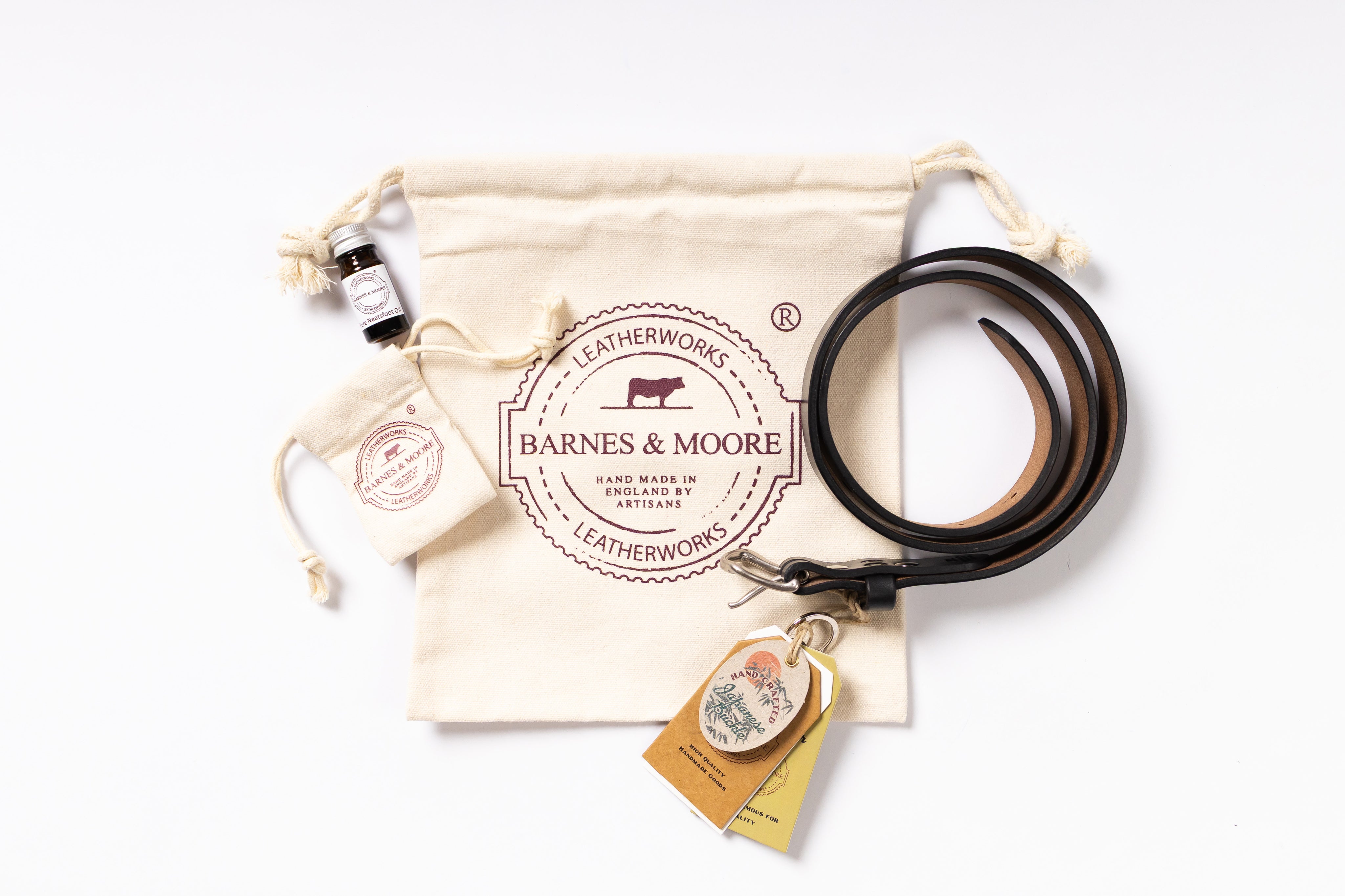 A leather belt presented with branded packaging on a white background, including a cream drawstring pouch, a small bottle, a branded tag, and a hanging leather tag. The belt is dark brown with a black edge.