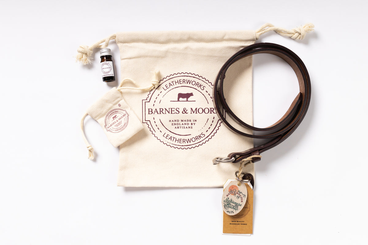 A leatherworking gift set from Barnes & Moor's on a clean white background, featuring a dark brown leather strap, a cream canvas drawstring pouch with a printed logo, a small dark bottle, a smaller matching pouch, and a hanging kraft paper tag.