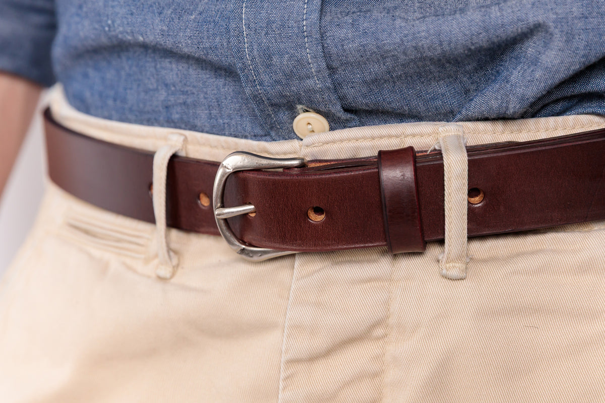 A close-up of a dark brown leather belt with a silver-tone buckle, worn over beige trousers and a blue denim shirt.