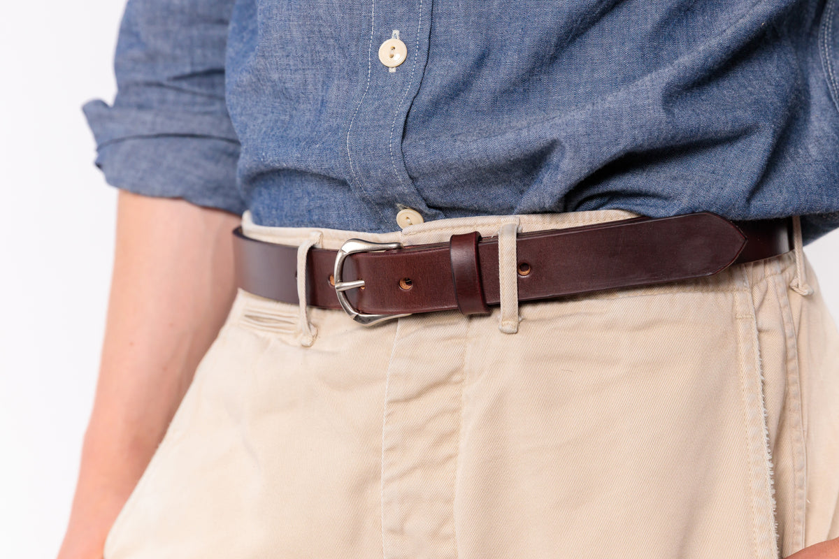 A close-up of a person wearing a dark brown leather belt with a silver buckle, tucked into beige trousers and paired with a blue button-down shirt.