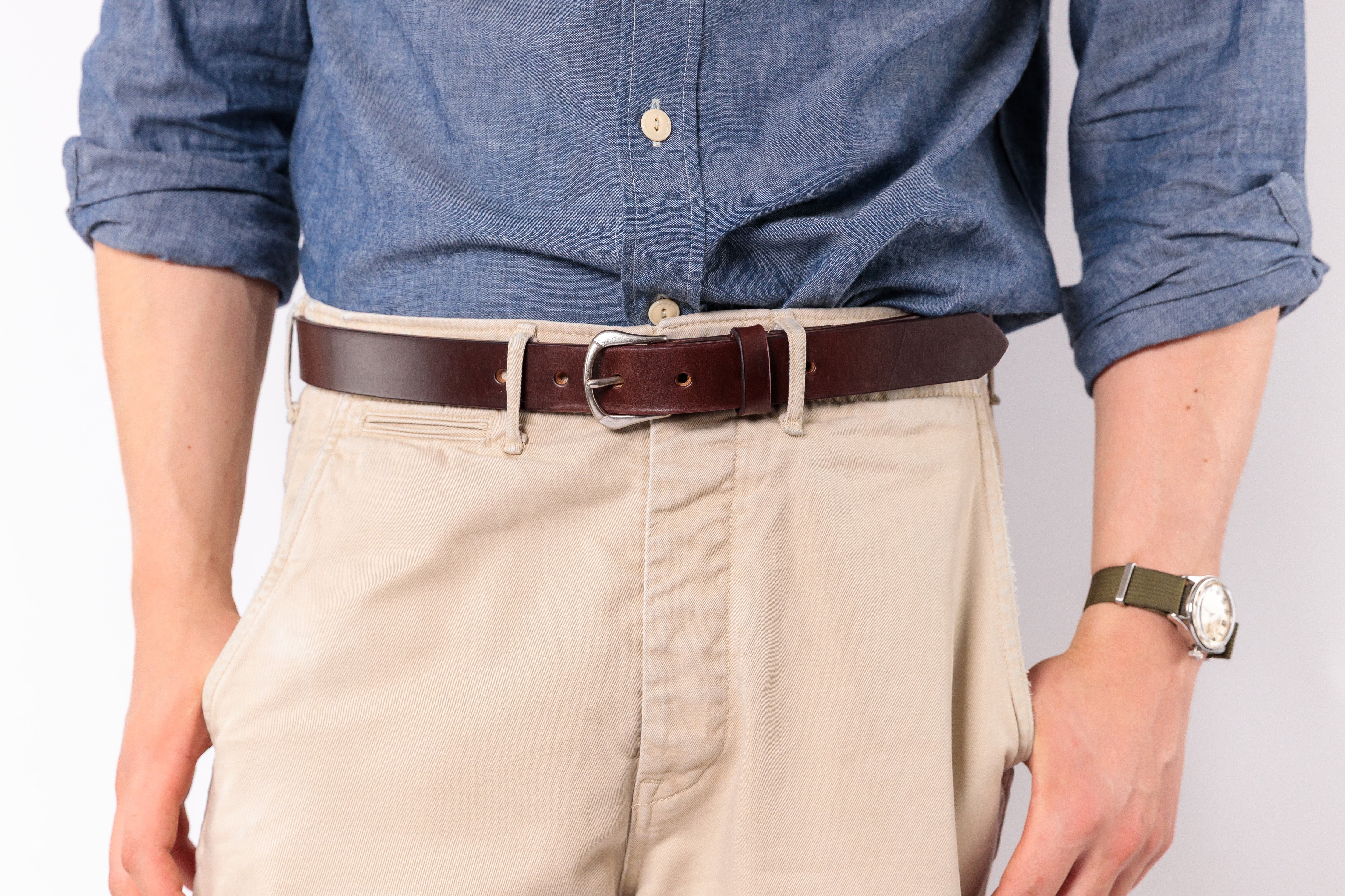A close-up of a person wearing a dark brown leather belt with a silver buckle, styled with beige chino pants and a blue chambray shirt.