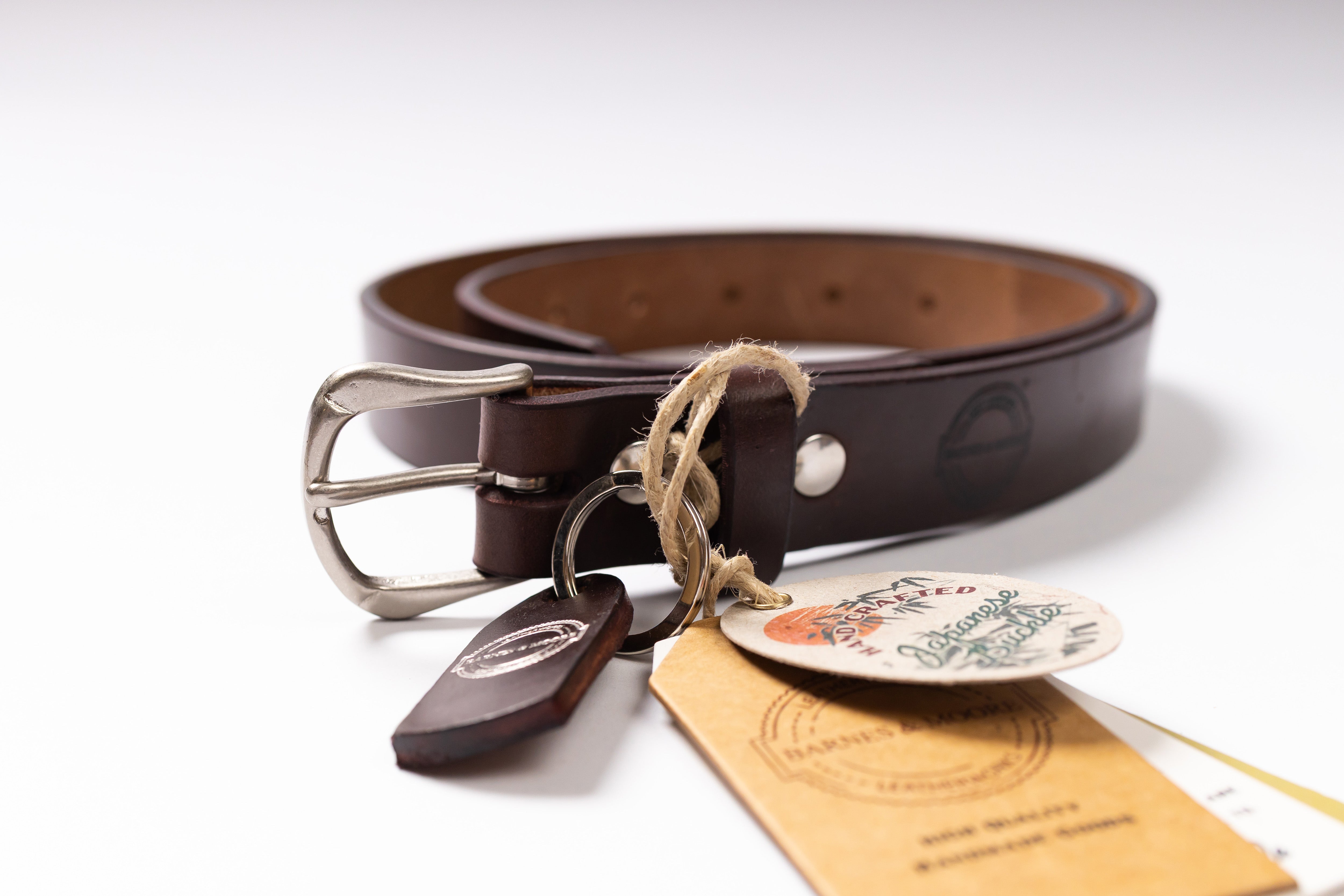 A dark brown leather belt with a silver metal buckle, shown coiled on a white background with its branded paper tags and wooden tag tied to the belt loop.