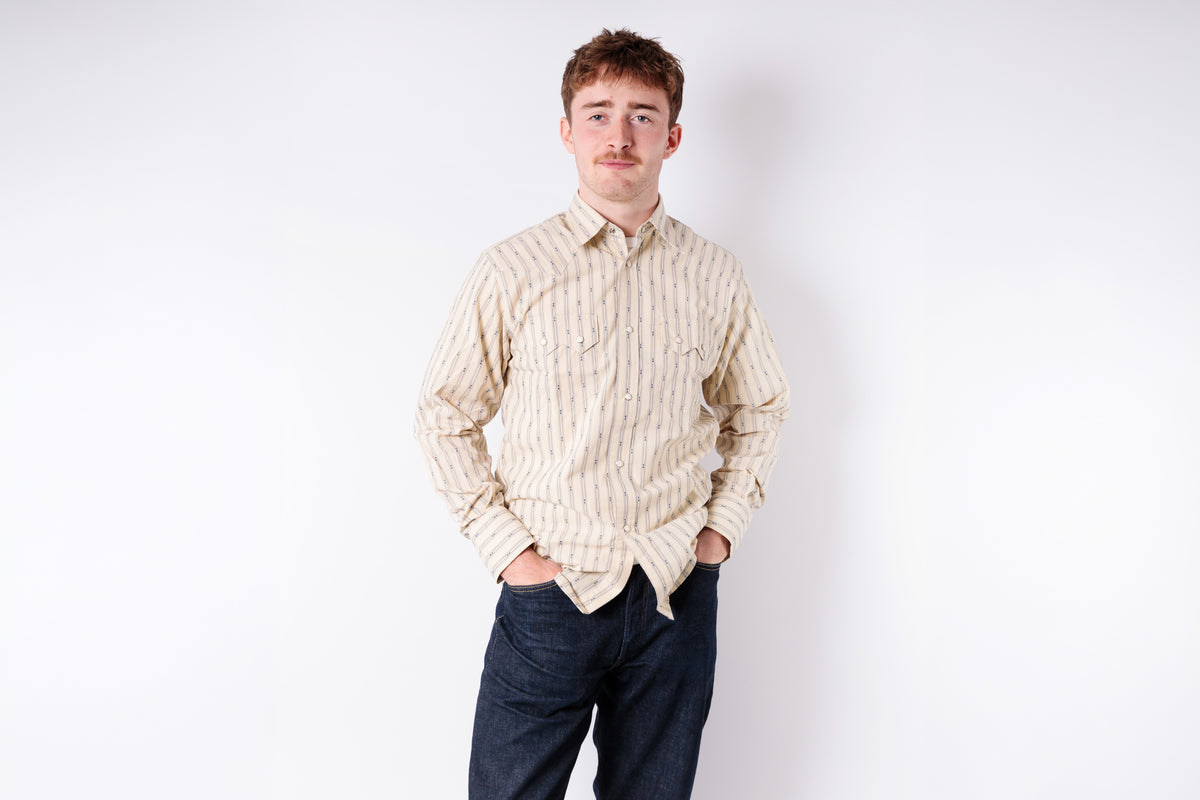 Man wearing a beige button-up shirt and dark pants on a white background