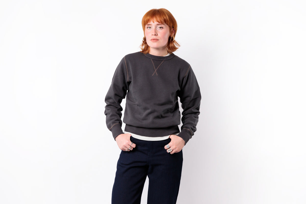 A person with red hair wearing a dark charcoal gray crewneck sweatshirt with a thin brown geometric line detail on the chest, paired with dark navy pants, standing against a plain white background.