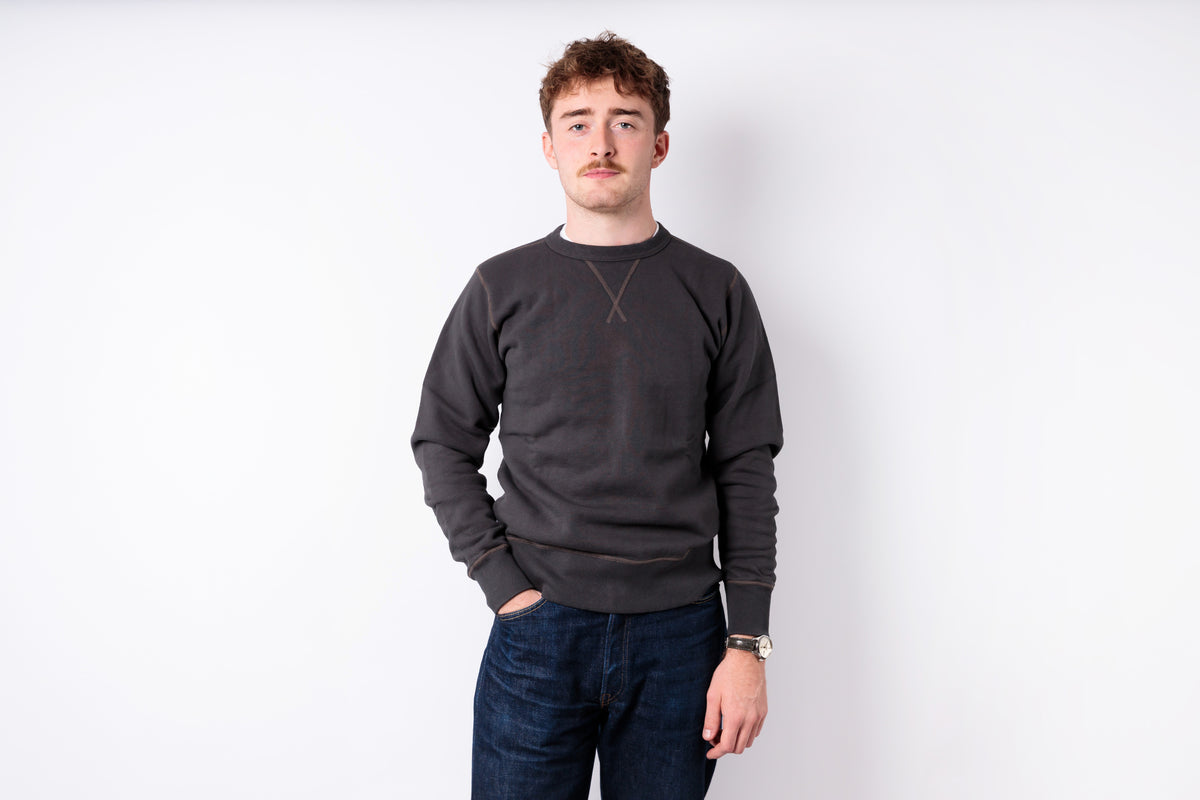 A man wearing a dark charcoal gray crewneck sweatshirt and dark blue jeans, standing against a plain white studio background.