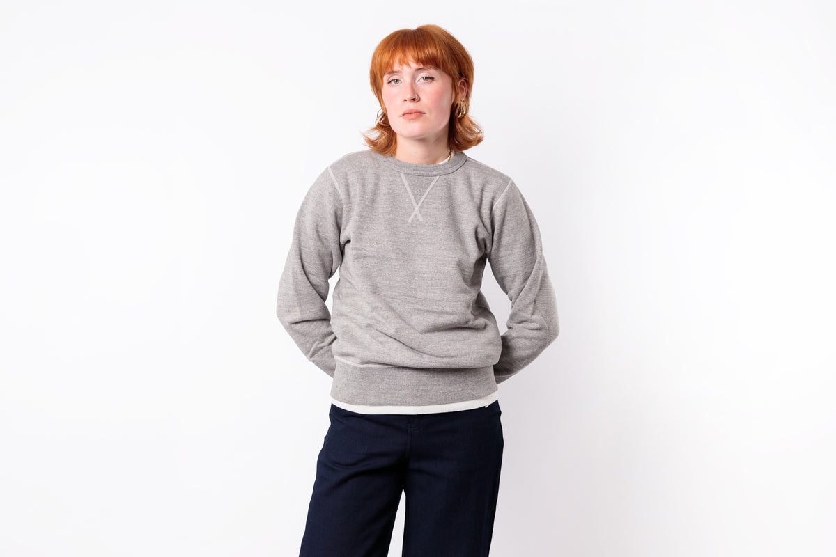 A woman with short red hair wearing a light gray knit crewneck sweater with a white geometric V-shaped chest detail and dark navy pants, standing against a plain white background.