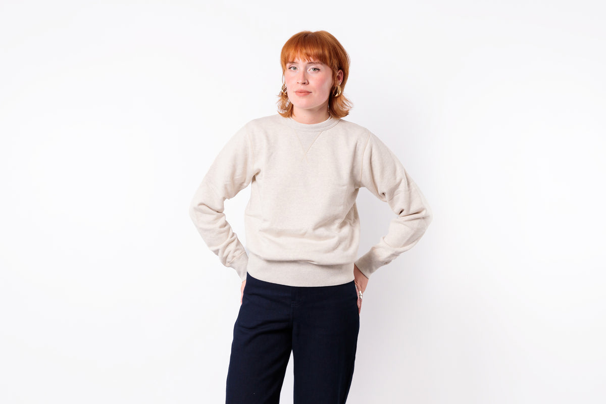 A woman with short red hair wearing a light beige crewneck sweatshirt and dark navy pants, standing against a solid white background.