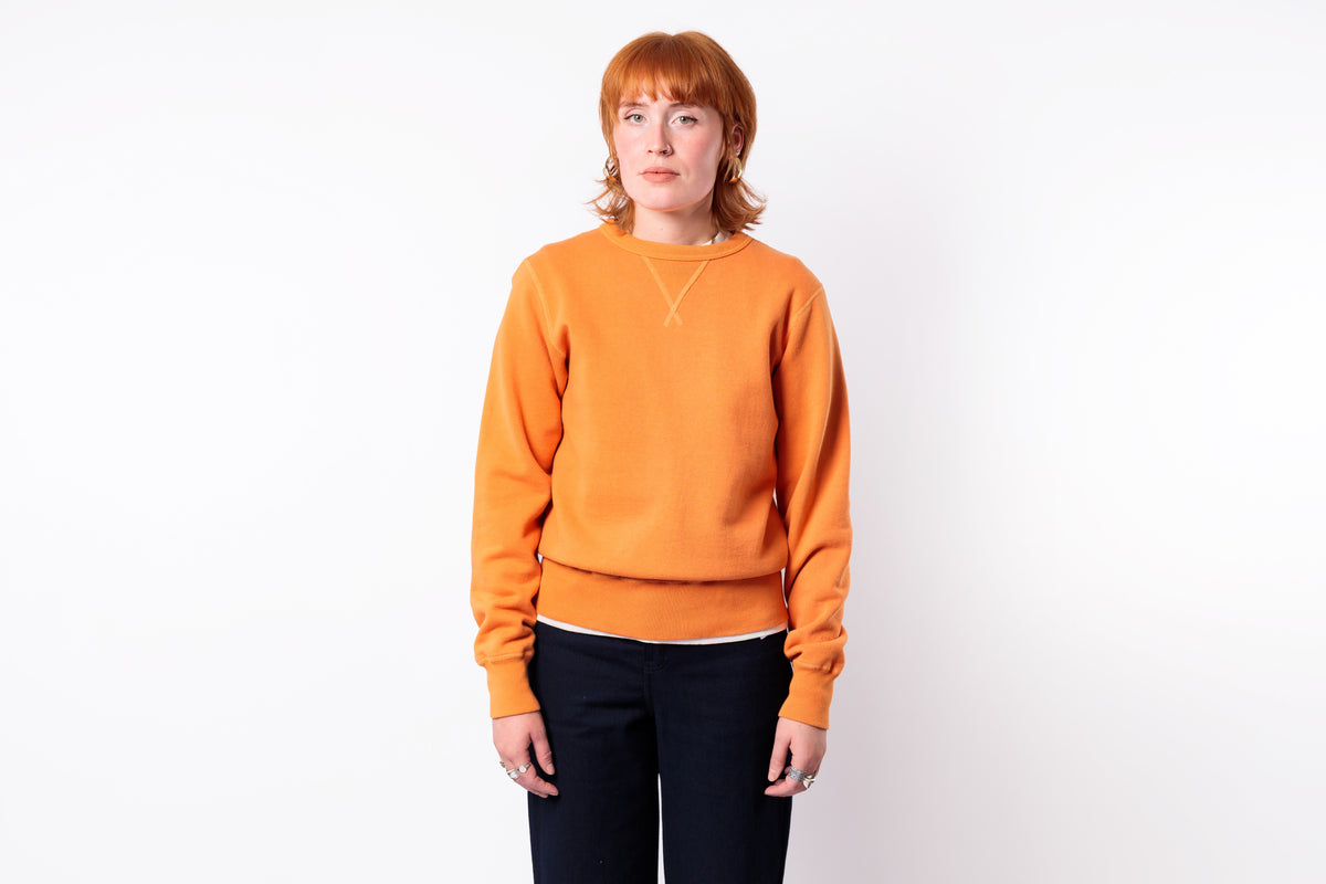 A woman with short red hair wearing a bright orange crewneck sweatshirt and dark navy pants, standing against a plain white studio background.