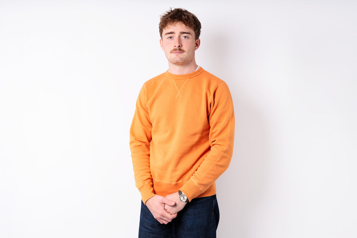 A young man with short brown hair and a mustache wearing a bright orange crewneck sweatshirt and dark blue jeans, standing against a plain white studio background.