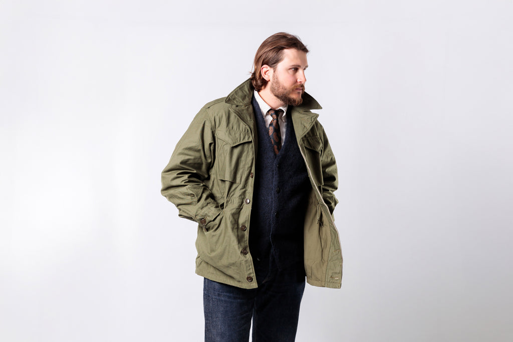 Man wearing a green jacket over a dark sweater and jeans on a white background